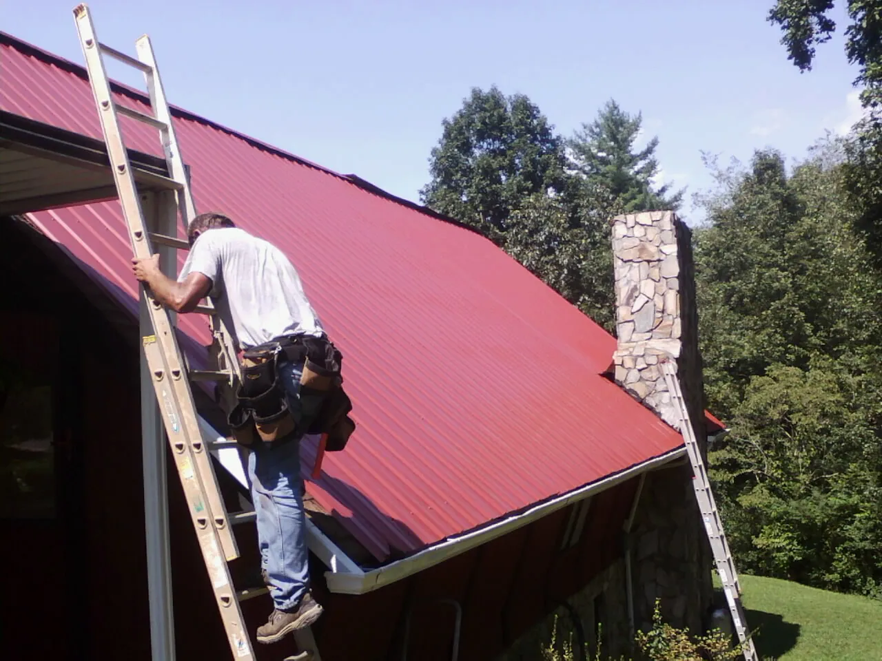 Roofer on a ladder beside a red metal roof and stone chimney — Soffit & Fascia Repair by Roofing Brothers in Mogadore